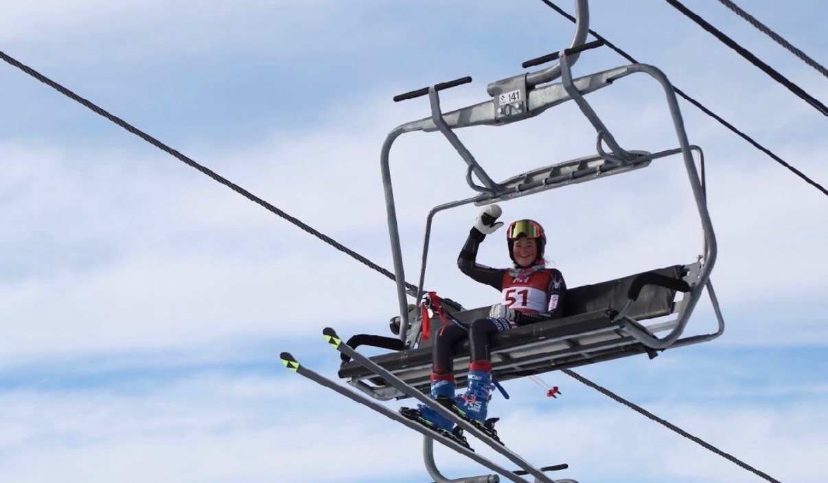 Bayli riding a chairlift in a red USSA race bib #51 with skis on